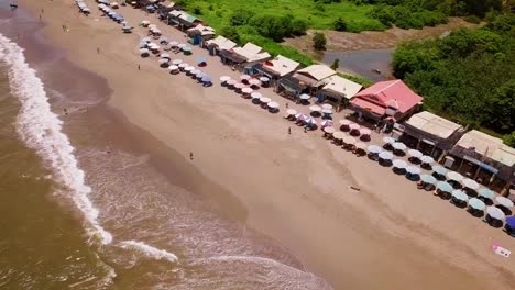 Aerial-View-Of-A-Beach-With-Beach-Bars,-Umbrellas,-Beach-Chairs-And-People-Walking-Along-The-Shore