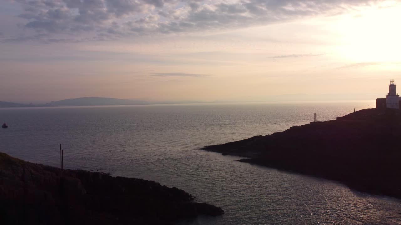 Espectacular toma aérea al amanecer del Faro de Mumbles en la isla, con una suave iluminación de hora dorada y la costa galesa de fondo. Ideal para turismo y documentales de viaje. Gales, Reino Unido. 4K.