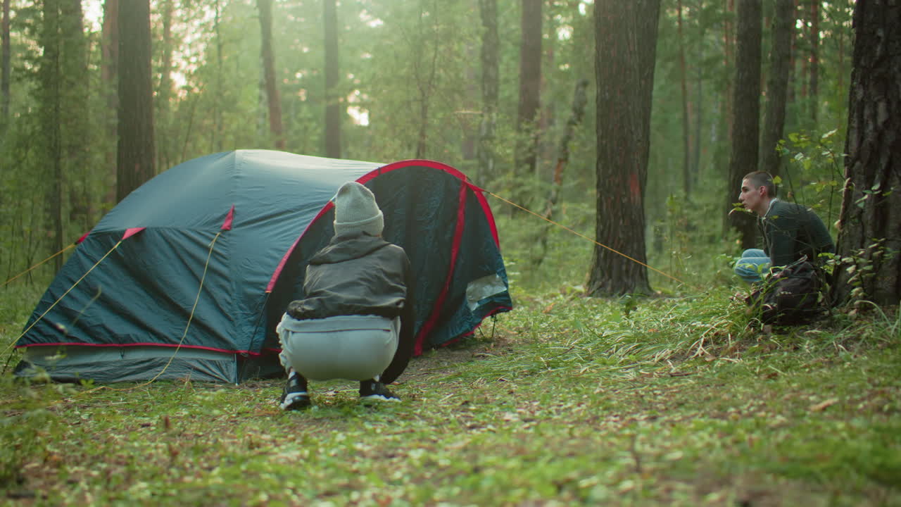 Two campers crouch on opposite sides of forest tent working to finalize setup, surrounded by tall trees and morning light filtering through woodland