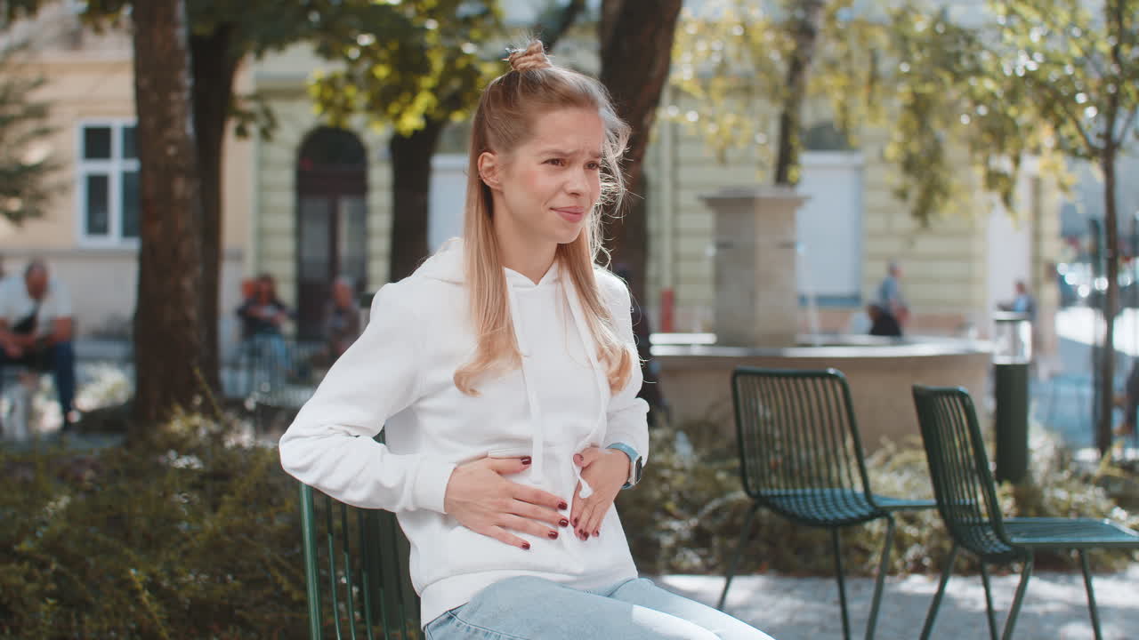 Sick ill young woman suffering from period cramps painful stomach ache sitting on chair in city