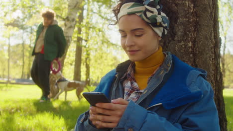 woman using phone in park and looking at playing dog