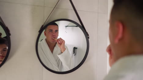 man brushes teeth looking at mirror near wife. guy in bathrobe carefully does hygienic routine standing near wife in bathroom. everyday care procedure