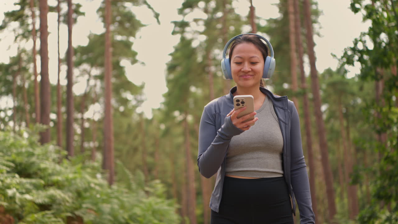 mujer con auriculares inalámbricos haciendo ejercicio haciendo ejercicio al aire libre transmitiendo música o podcast desde un teléfono móvil corriendo por una pista en el bosque con ropa deportiva filmada en tiempo real 1