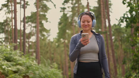 mujer con auriculares inalámbricos haciendo ejercicio haciendo ejercicio al aire libre transmitiendo música o podcast desde un teléfono móvil corriendo por una pista en el bosque con ropa deportiva filmada en tiempo real 1