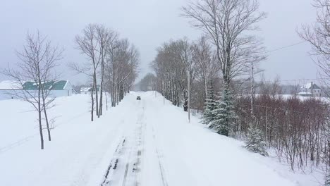 flying down a snow covered tree lined road during a blizzard toward a black car slow motion aerial