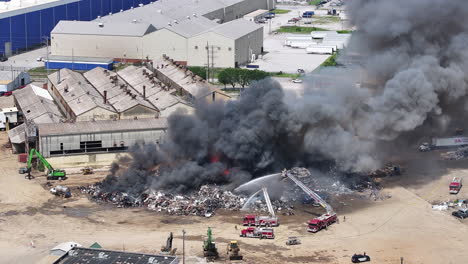 slow motion aerial footage of a large scrap metal fire with emergency services covering it with water as black smoke moves into the sky