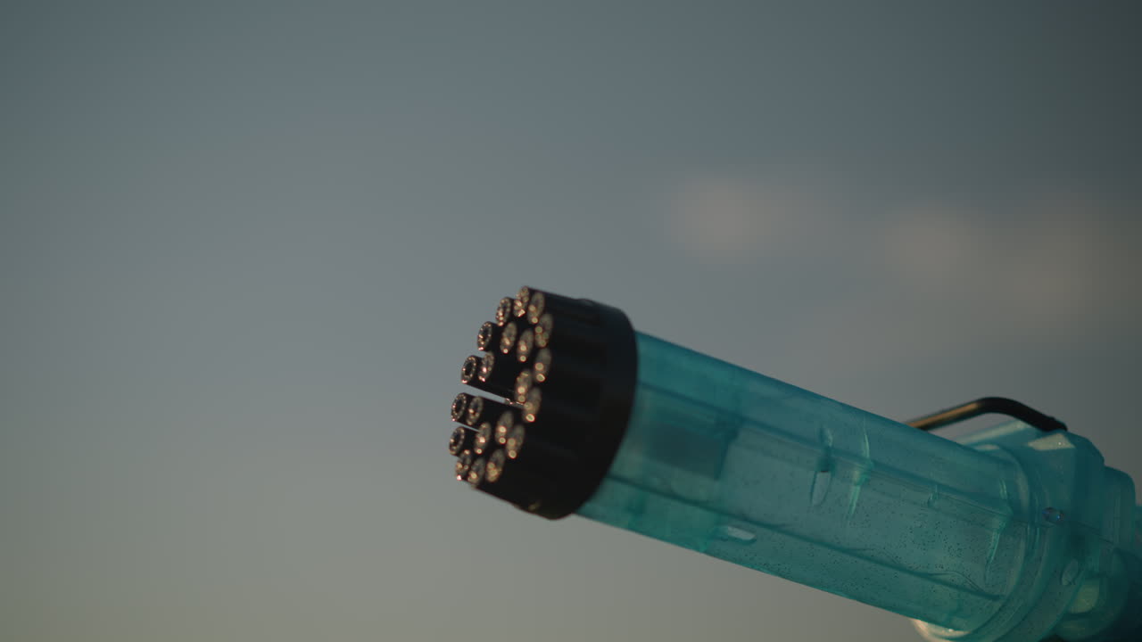 Close-up of a plastic bubble gun releasing a stream of colorful bubbles under a clear sky. The vibrant bubbles catch the light, creating a magical and playful scene