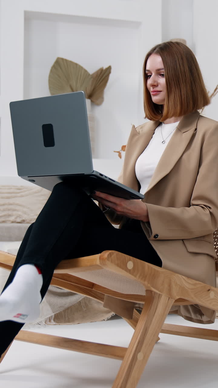 Business lady in beige jacket sits in armchair. Woman opens her laptop, puts on glasses and starts her work. Vertical video
