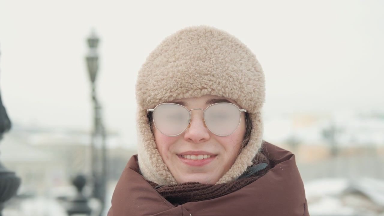 Ritratto frontale testa e spalle di un adulto che indossa un cappello da trapper in maglia con paraorecchie, occhiali trasparenti, una giacca imbottita con cappuccio e una sciarpa di maglia mentre si trova all'aperto in un parco urbano innevato, calmo e freddo
