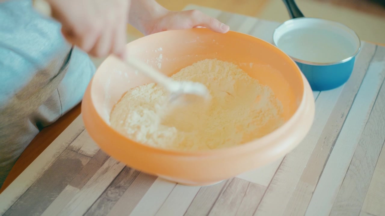 Close up of teenage hands mixing dry and liquid ingredients with a wooden spoon in an orange bowl. Starting homemade bread dough, cozy kitchen lifestyle, authentic learning process, 4k.