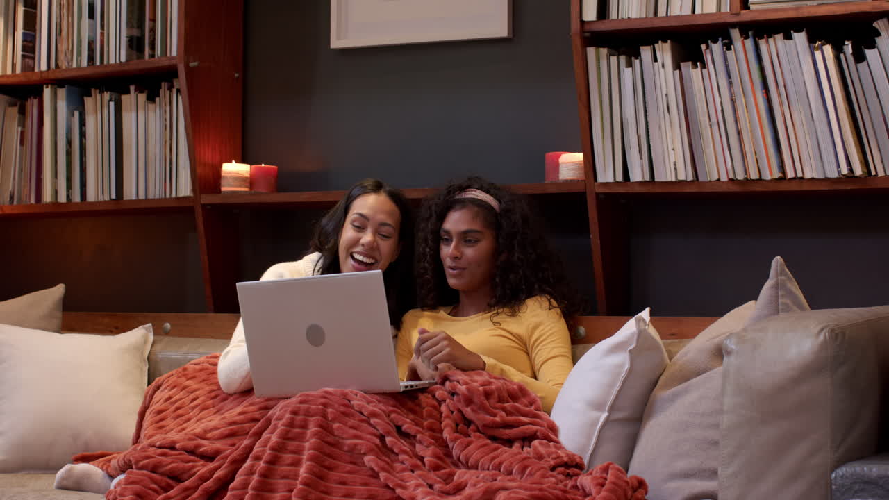 Two multiracial female friends relaxing on couch with laptop, enjoying cozy evening, at home