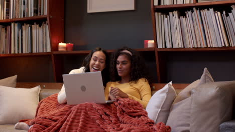 two multiracial female friends relaxing on couch with laptop, enjoying cozy evening, at home