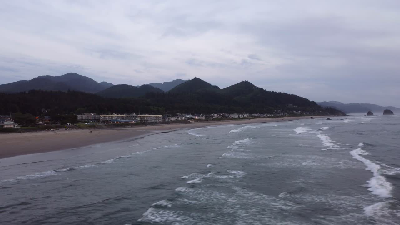 Aerial drone shot of rows of waves crashing onto Cannon beach in Oregon during sunset. Hotels and lush green hills tower over the beach and the forest contrasts the blue ocean and sandy beach.