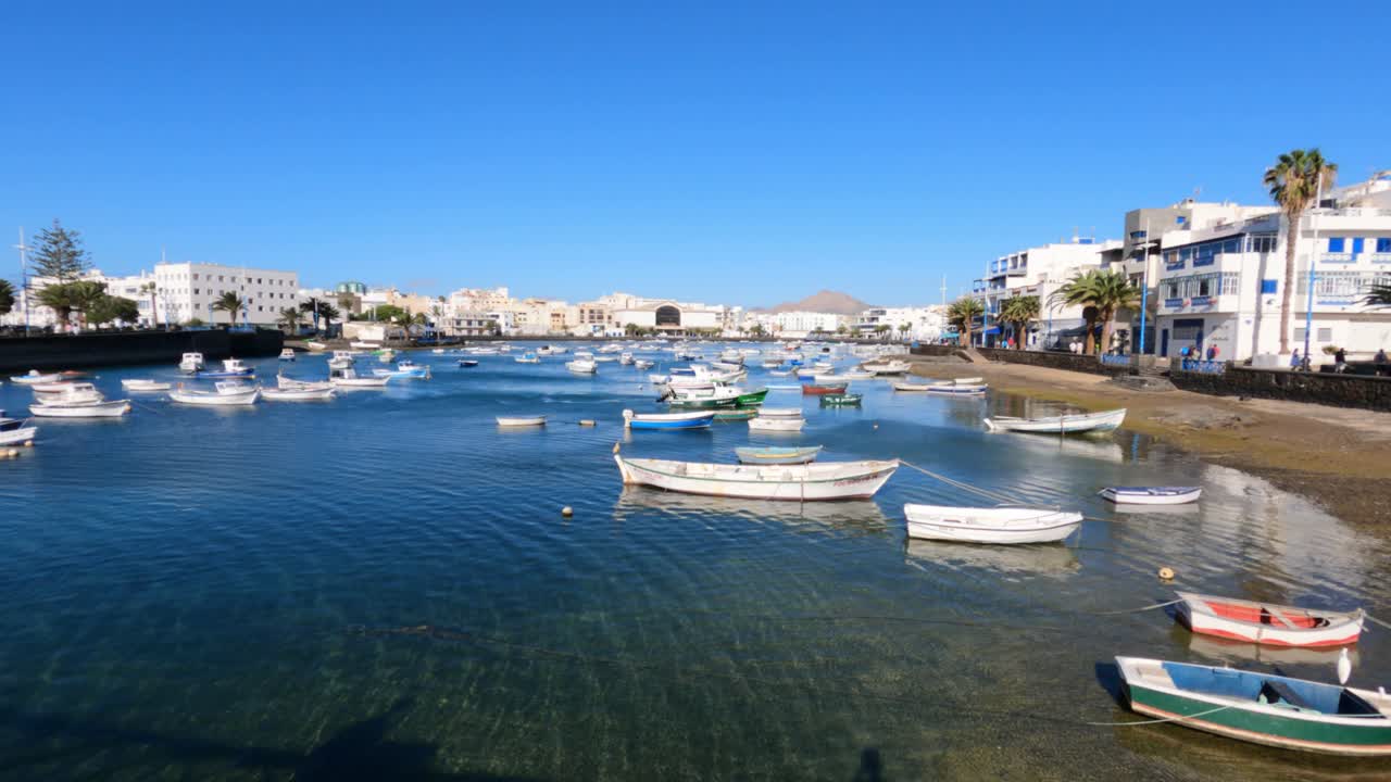 Scena portuale tranquilla ad Arrecife, Lanzarote, con piccole barche da pesca ancorate nella serena laguna del Charco de San Ginés sotto un cielo azzurro vivido.