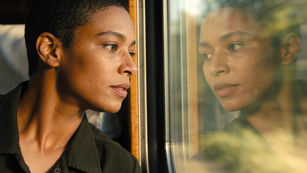 Leaning woman in shirt on train with sunlight hitting face, shifting gaze to reflection on glass