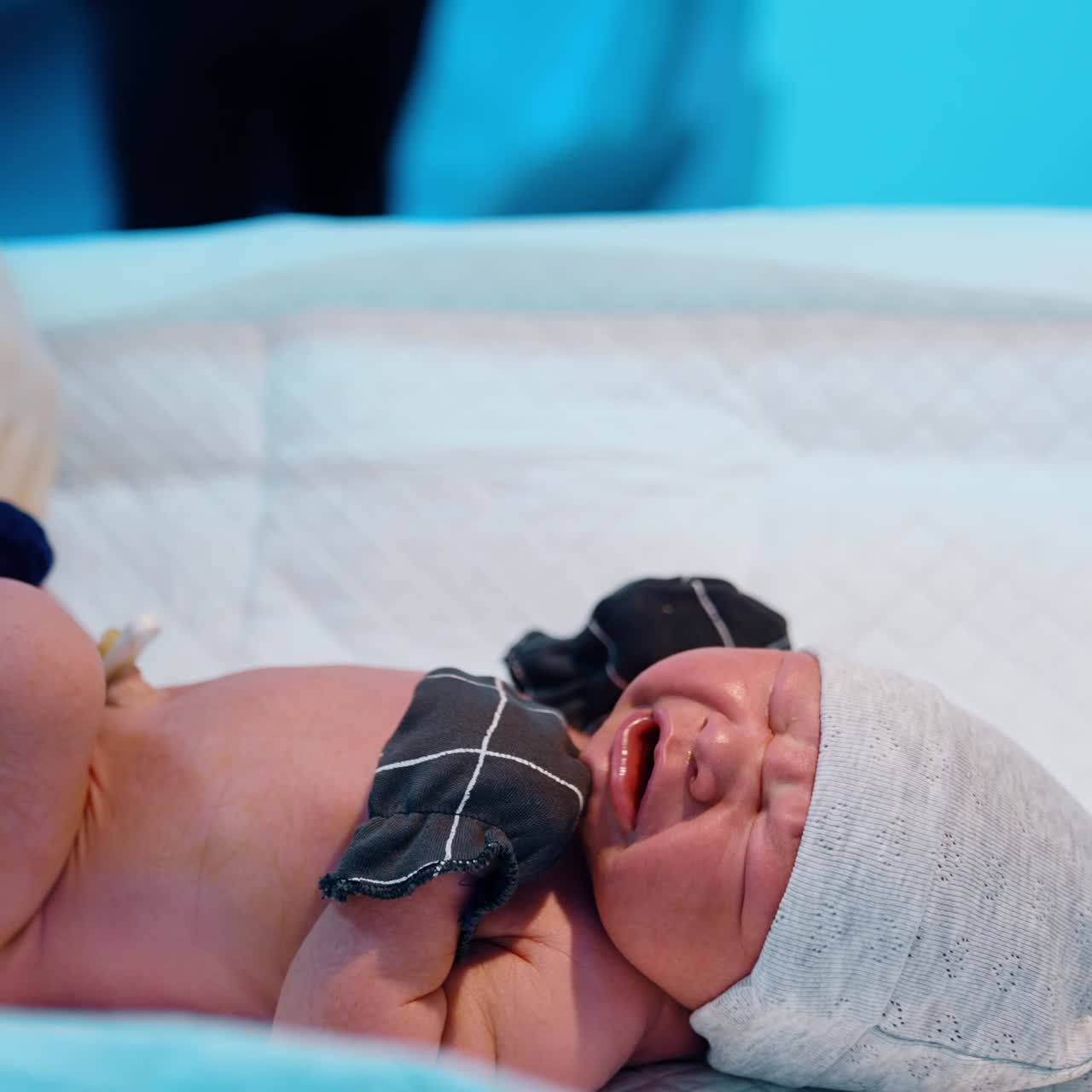 Newborn wearing mittens, socks and cap lies on the table crying. Nurse outs on a diaper on a baby