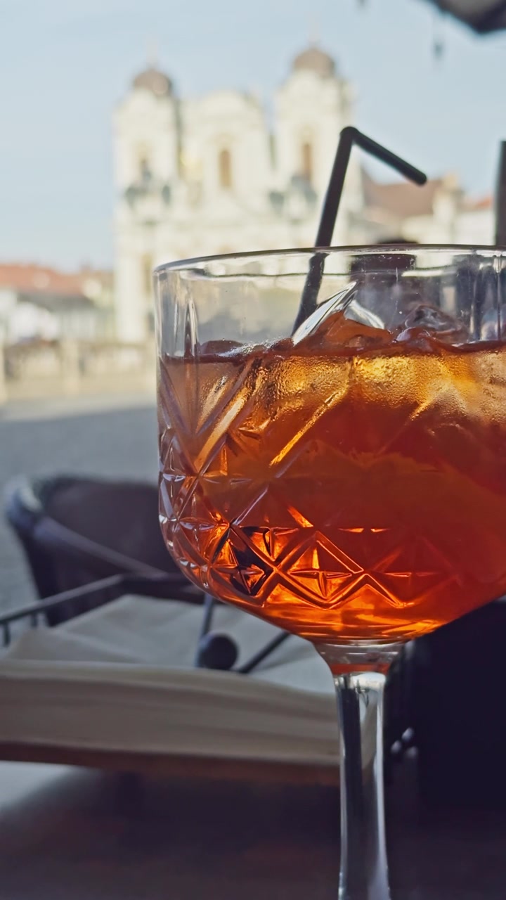 Primo piano di un rinfrescante Aperol Spritz su un tavolo di un ristorante all'aperto con la Cattedrale di San Giorgio sullo sfondo, che cattura l'atmosfera della ristorazione europea e dei viaggi estivi a Timisoara
