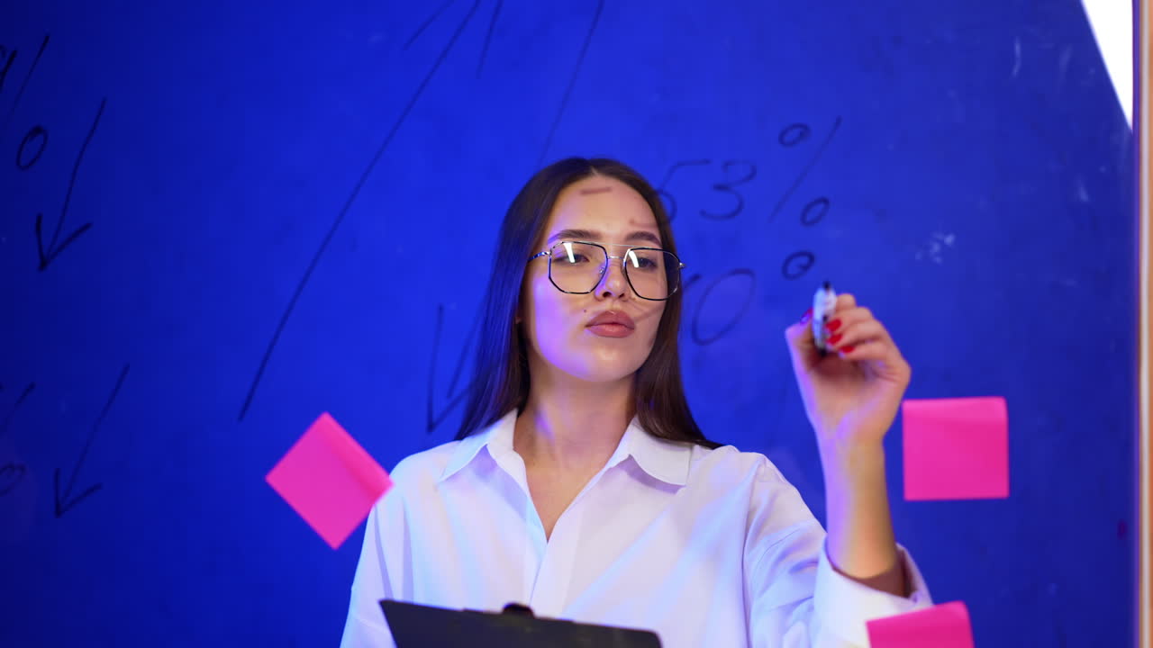 Good-looking lady in glasses and white shirt comes up to a glass board. Focused woman holding a clipboard writes with a marker. Business concept.