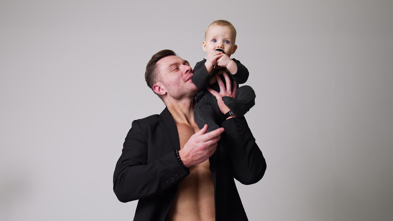 Handsome man wearing a black jacket on the naked torso holding a baby on his shoulder. Father and infant son portrait. Studio footage. White backdrop.