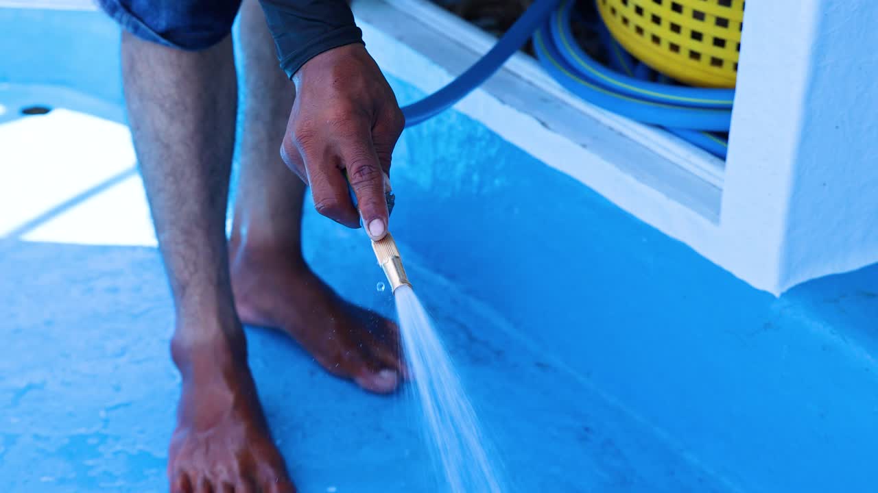 A person washes a blue boat deck with a hose, under bright daylight, in Phuket