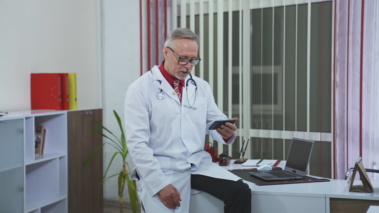 Medical consultation during quarantine. Elderly doctor in glasses sitting in his cabinet and having video call to a patient using phone.