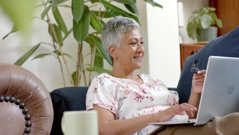 Happy-senior-biracial-woman-sitting-on-couch-and-using-laptop-at-home,-slow-motion