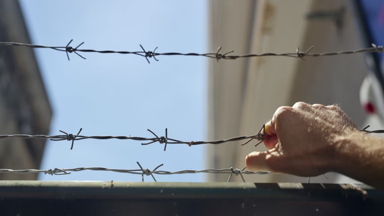 Hand approaching barbed wire on bright day. Sharp metal strands against blue sky. danger, restriction, borders, security, imprisonment, migration, or human-rights themes. Locked or imprisoned. hope