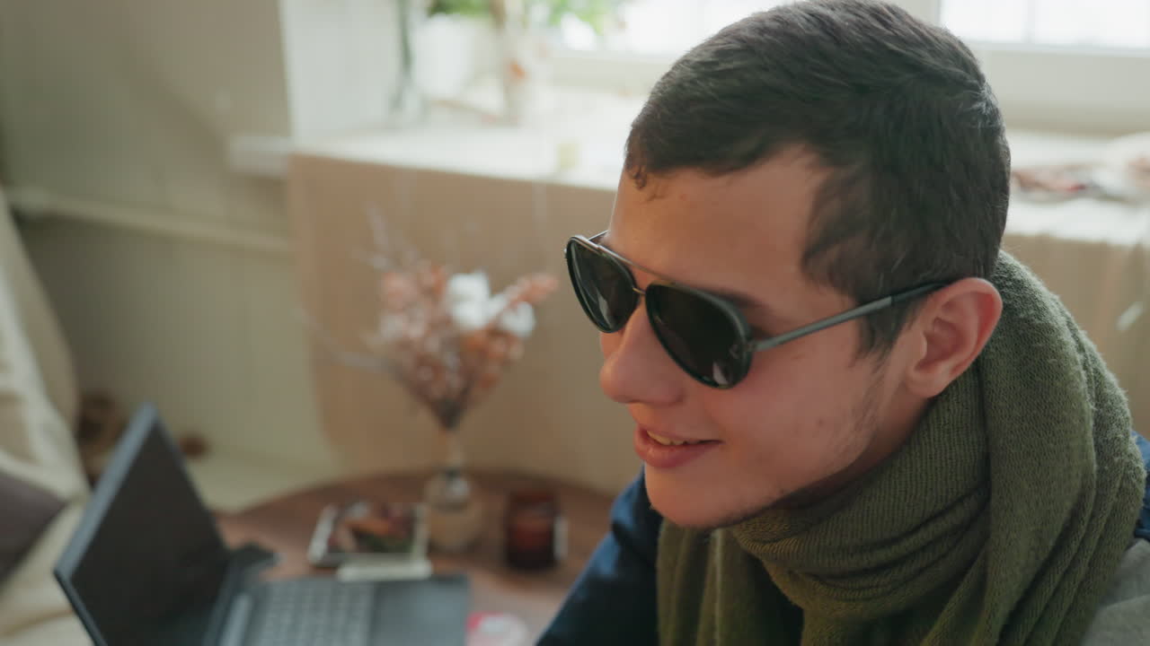 Young man indoors trying on sunglasses in cozy apartment with laptop and green scarf, soft window light and warm bokeh, contemplative expression capturing freelance workspace mood