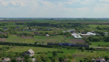 rural commute in an old blue train, travelling between houses and green fields on a sunny clear day, aerial tracking shot