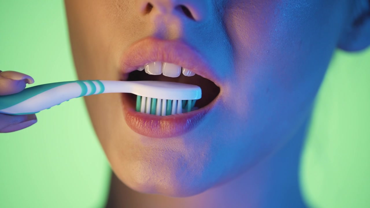 Slow motion 4K macro shot of a woman brushing her teeth with a toothbrush, vibrant green background with glowing pink and blue neon light on her face, cinematic and clean, detailed close-up aesthetic