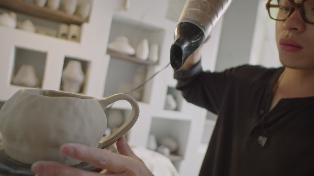 Young Asian Ceramist Drying Pottery with Hairdryer