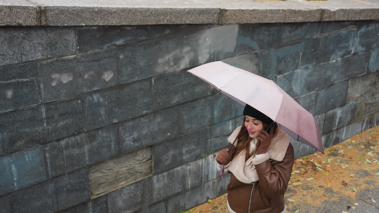 Up shot of young girl smiling on phone call holding umbrella, wearing knit cap, brown shearling jacket, black trousers, walking down stone steps during light snowfall with colorful autumn leaves