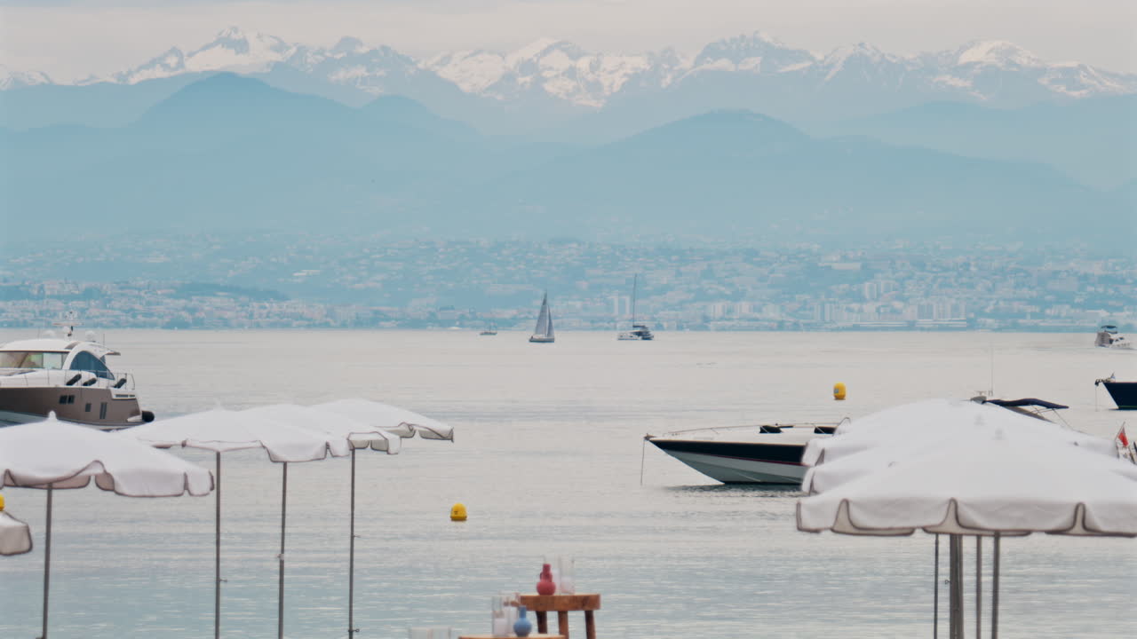 Close up of white sun umbrellas on the beach with multiple boats on the sea, the city and mountains on the background