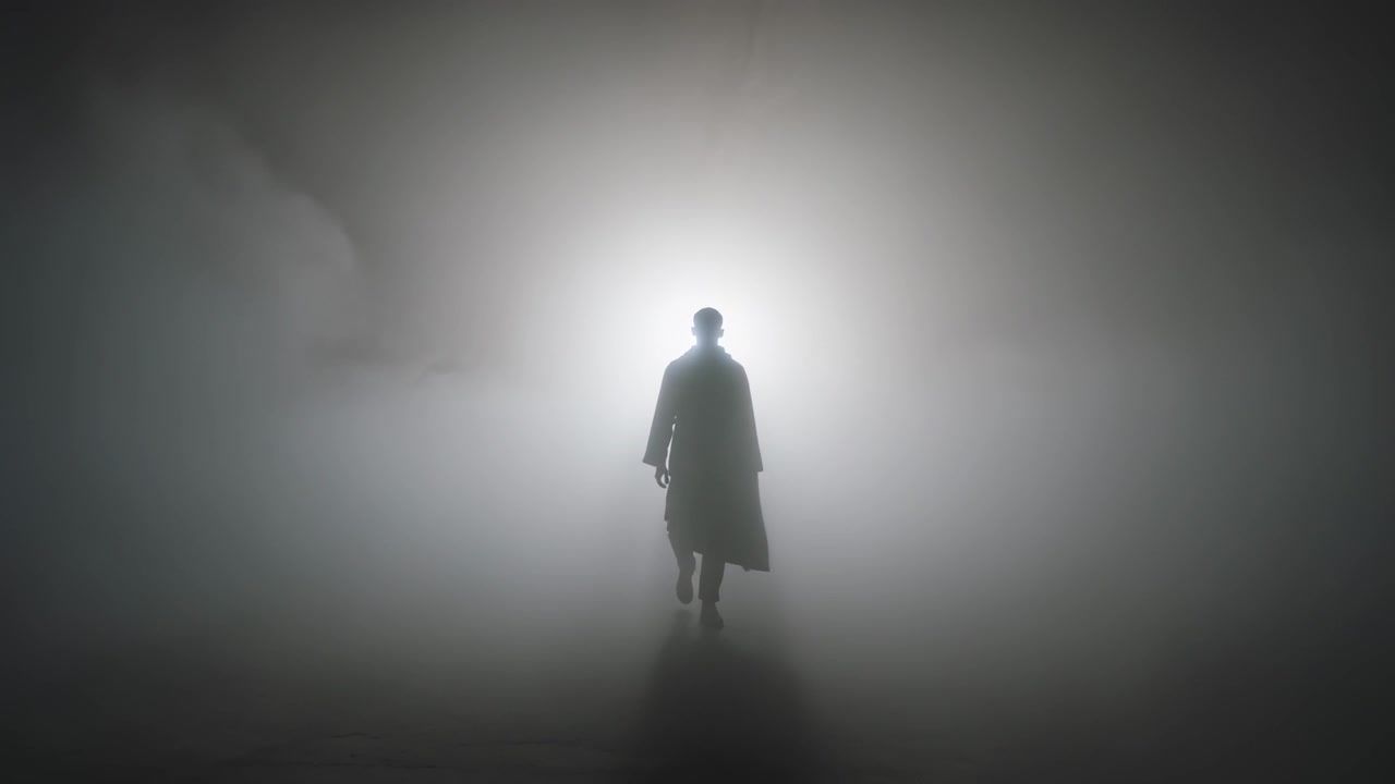Walking man stepping from bright halo toward camera in fog, wearing long coat and brimmed hat