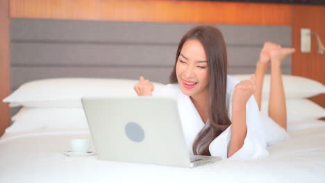 a young woman in a terry cloth robe, lying on her stomach on a comfortable hotel bed works on her laptop