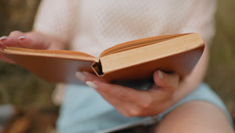 primer plano de manos sosteniendo un libro abierto, hojeando páginas, borrón suave en el fondo, con pantalones cortos azules y un momento de lectura relajado en un entorno tranquilo al aire libre