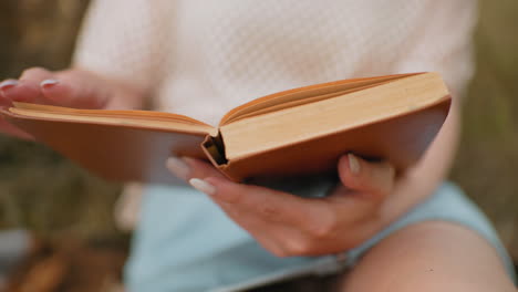 close-up of hands holding an open book, flipping through pages, soft blur in background, featuring blue shorts and a relaxed reading moment in a peaceful outdoor setting