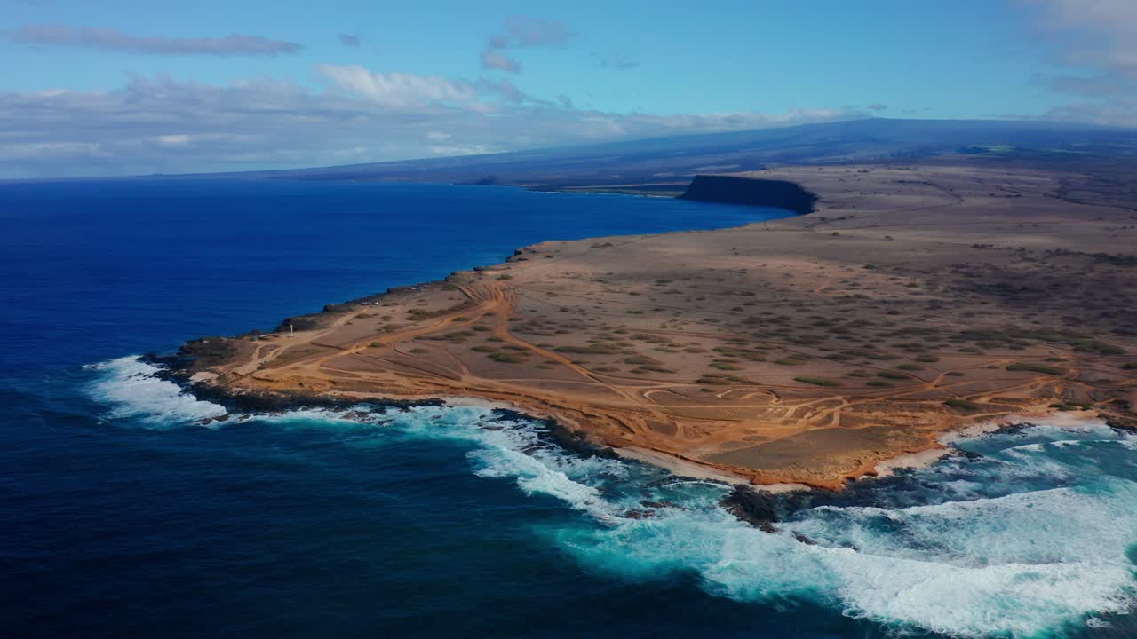Rough blue surf crashes against a sweeping rocky shore lined with winding dusty trails, all under a vast open sky stretching to the horizon.