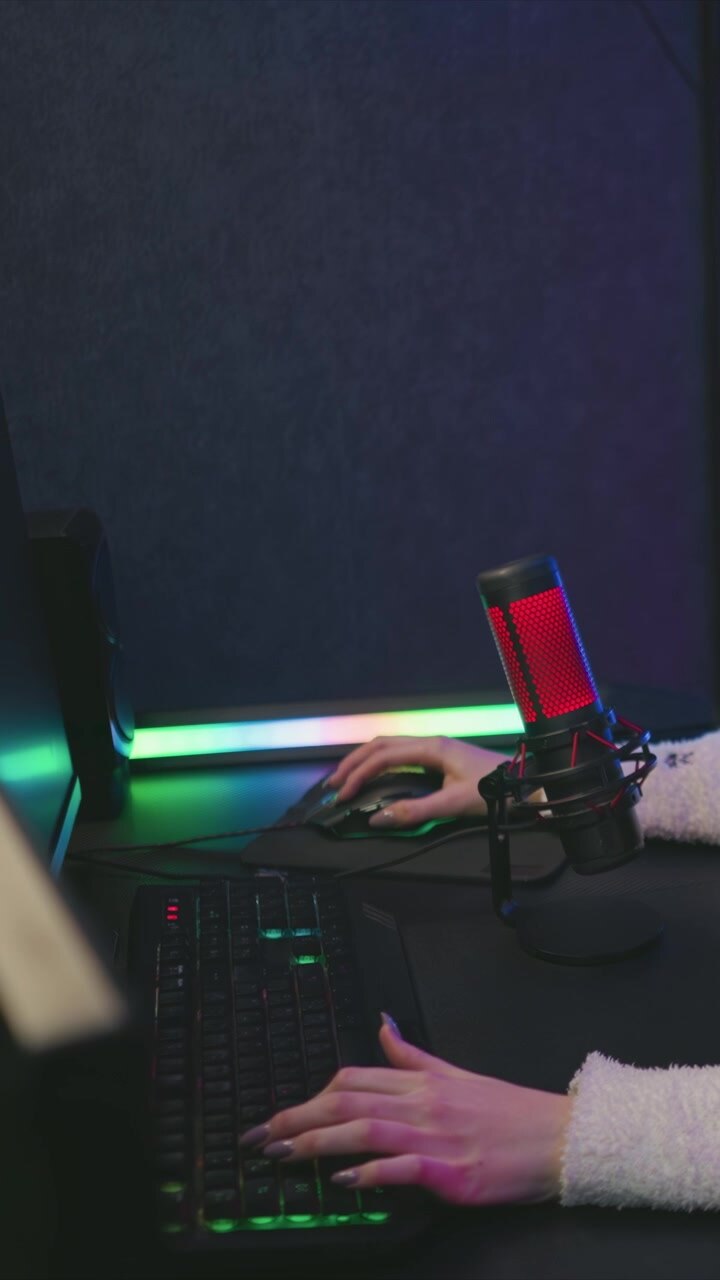 Giovane donna digita sulla tastiera e usa il mouse alla scrivania con un'illuminazione LED colorata, indossando una manica bianca soffice. Un microfono rosso è posizionato vicino al monitor in un ambiente studio interno con luce soffusa