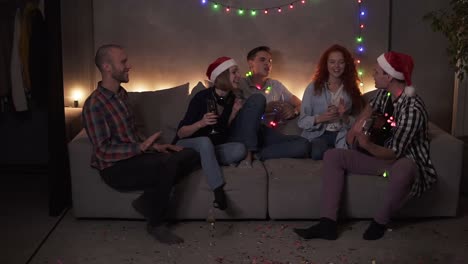 amigos cercanos alegres celebrando, pasando tiempo juntos en casa. amigos felices se sientan en el sofá y escuchan a un chico cantando y tocando la guitarra. reunirse en navidad. luz apagada, luces de guirnalda. camara lenta