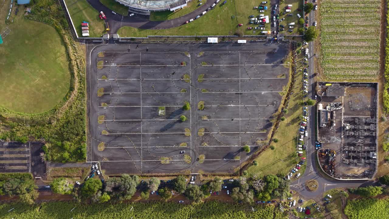 Aerial top-down view of an empty car park near a racing event area in Mauritius, surrounded by green fields and stadium grounds, captured by drone in UHD quality