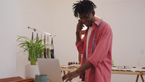 happy african american craftsman with dreadlocks making phone call and using laptop in workshop