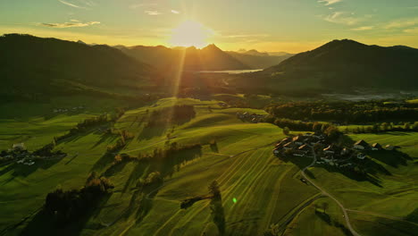 Aerial-shot-capturing-the-sunrise-shining-over-the-green-grassland-with-trees-and-houses-in-the-distance