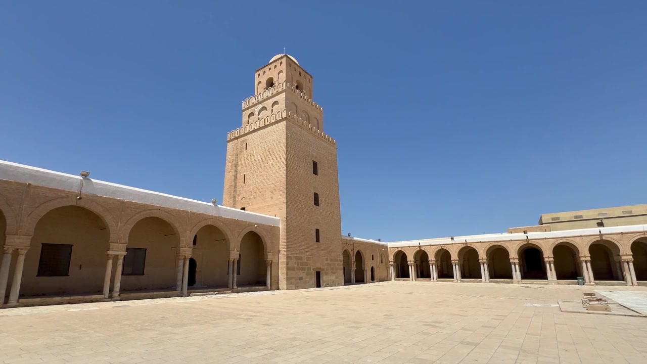 Bredt udsyn i dagtimerne over Den Store Moské i Kairouans gårdhave med dens høje murstensminaret og omkringliggende arkader, velegnet til historie- eller arkitekturindhold. Tunesien.