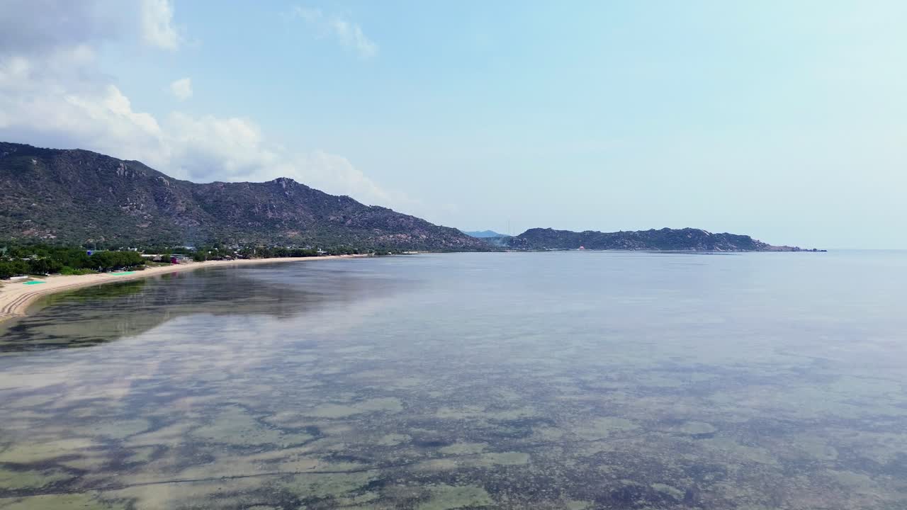 Drone flies forward and upward above calm water, with the beach on the left side of the frame, slowly rising into the sky on a bright, clear day in Vietnam.