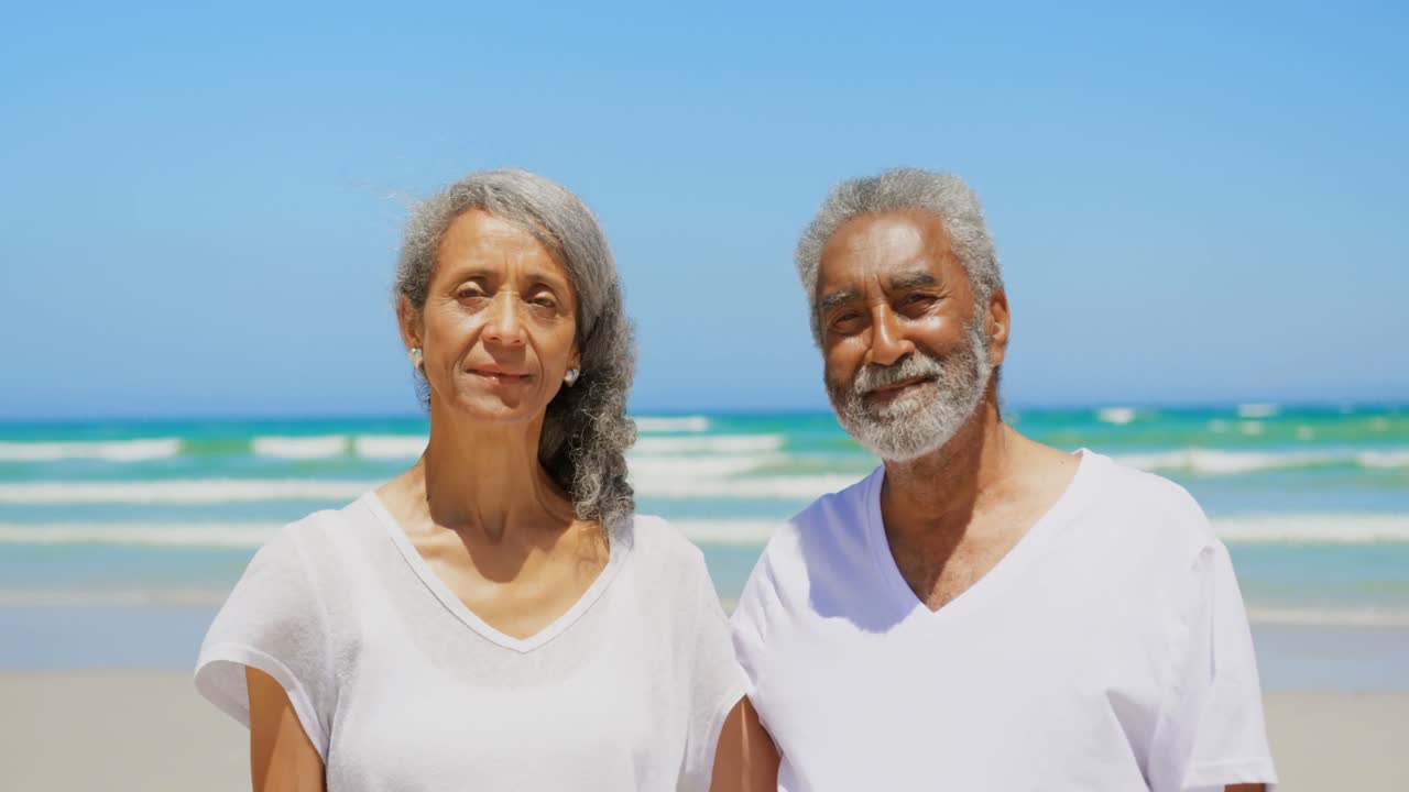 Front view of active senior African American couple with arms around standing on the beach 4k