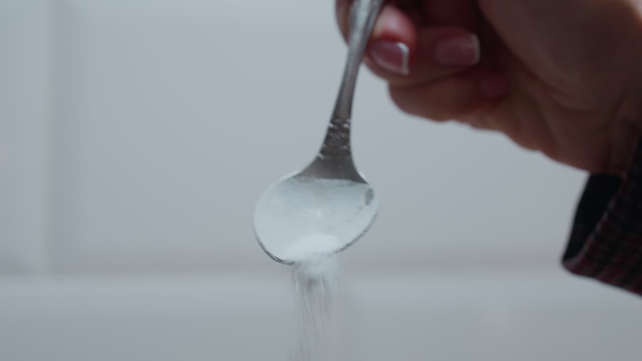 primer plano de la cuchara en la mano mientras el azúcar se vierte gradualmente con un fondo azulejoso brillante, las partículas finas de azúcar caen delicadamente, creando un momento culinario suave en un entorno de cocina bien iluminado