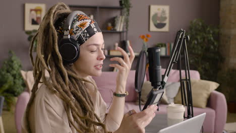 vista lateral de una mujer con rastas grabando un podcast con auriculares, hablando con un micrófono y leyendo en un smartphone sentado en un escritorio