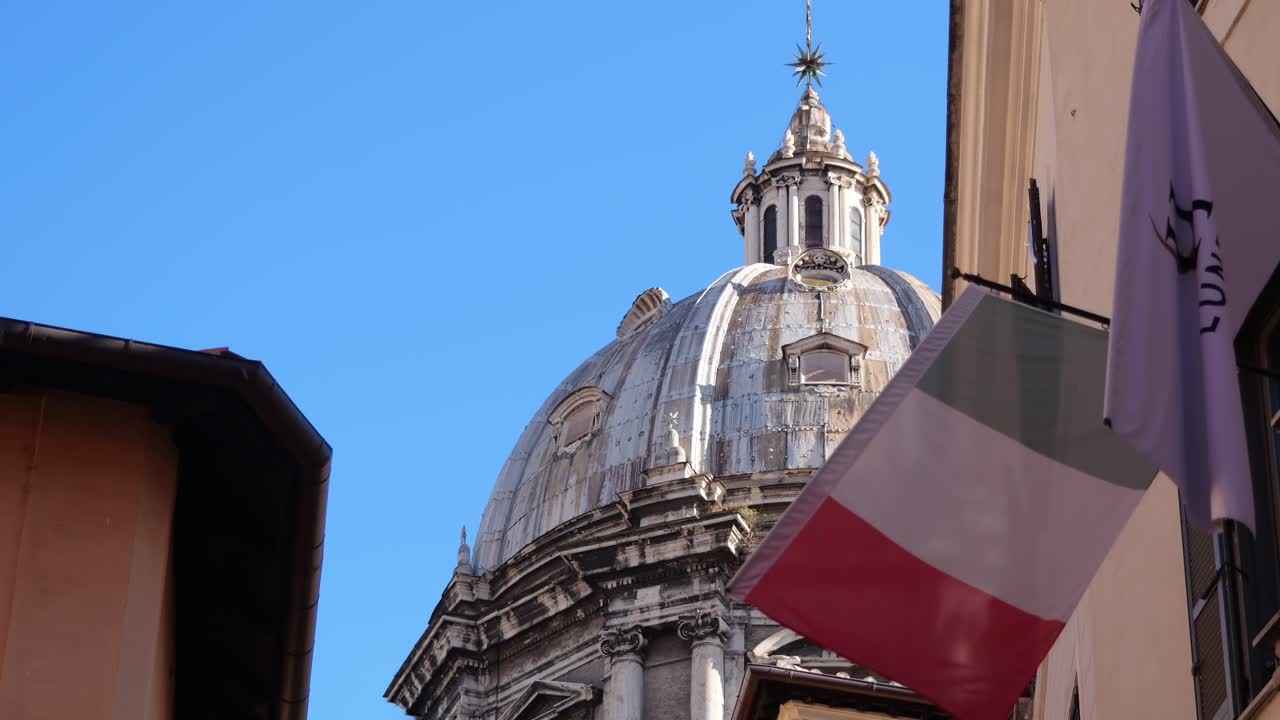 The Italian flag waves in front of an aged historic dome in Rome, Italy, surrounded by classic architecture and clear blue skies in a charming urban street setting.