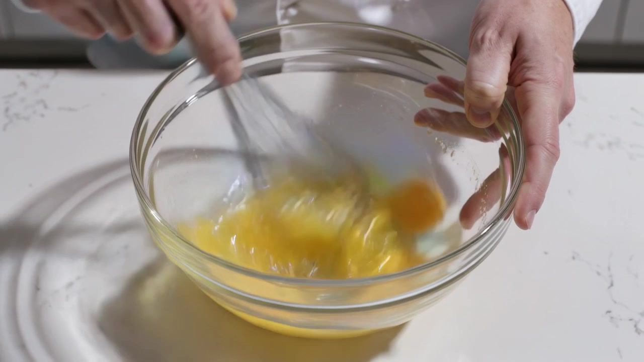 Placing whisk, hands in white sleeves whisking two yolks in glass bowl in kitchen for cooking